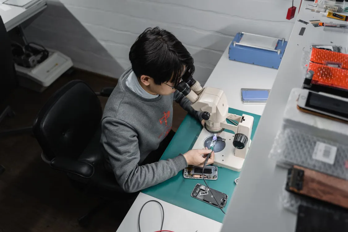 A technician examines an open smartphone under a microscope at a workshop bench.